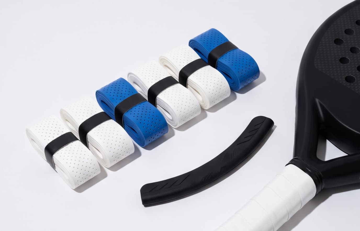 El Kit Grippada desplegado: seis overgrips perforados blancos y azules en fila, el protector de canto negro al lado y una pala de pádel negra con el grip blanco recién puesto.